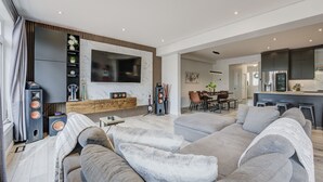 A modern living room with a grey sofa set, a flat-screen TV mounted on a marble wall, a wooden console, and a dining area with a table and chairs.