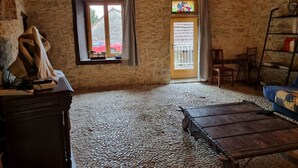 Ein rustikaler Raum mit Steinwand, Holzboden, einem Fenster mit Vorhängen, einer Tür mit Metalleinschlag, einem kleinen Tisch, einem Stuhl und einem Bücherregal.