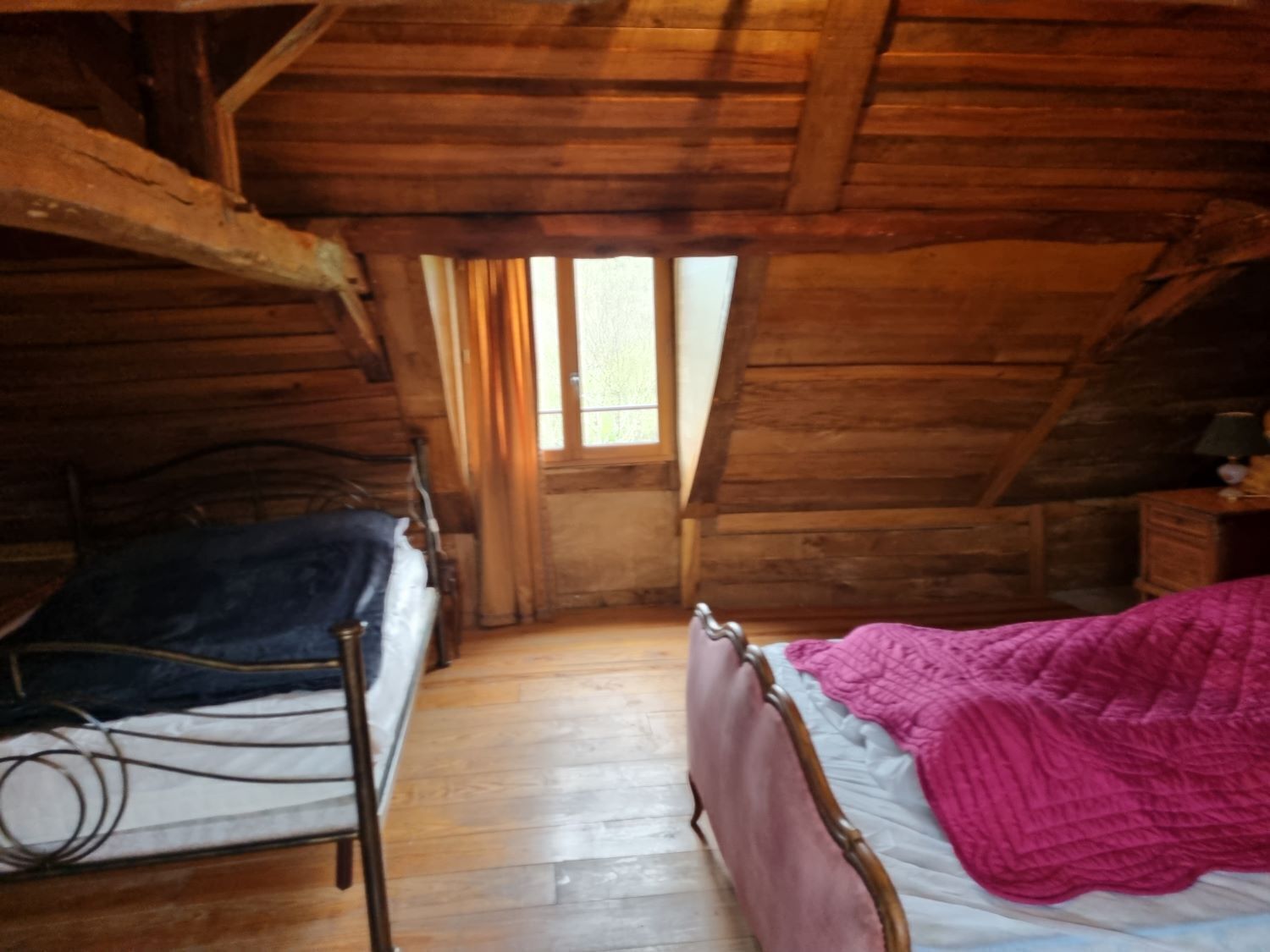 A wooden room with a sloped ceiling, a bed with a pink blanket, a bedside table, and a window.