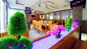 Un intérieur de restaurant moderne, avec des meubles en bois, un long comptoir agrémenté de plantes décoratives et un téléviseur fixé au mur.
