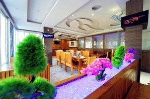 Un interior de restaurante moderno con mobiliario de madera, una larga barra con plantas decorativas y un televisor en la pared.