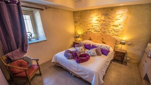 Un dormitorio con pared de piedra, una cama con cabecera, una silla de madera, un ventanal con vistas y una mesita de noche con lámpara.