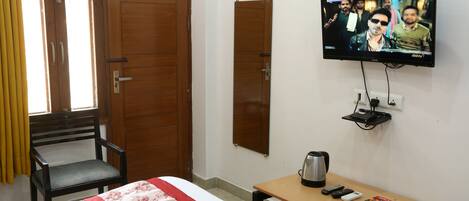 A hotel room with a bed, a wooden table with a kettle, a television, and a chair.