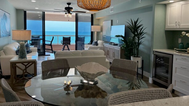 Een moderne woonkamer met een glazen tafel, rieten stoelen, een plafondventilator en uitzicht op de oceaan.