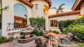 A patio with a stone fountain, outdoor seating, and a view of a house with a tiled roof and large windows.