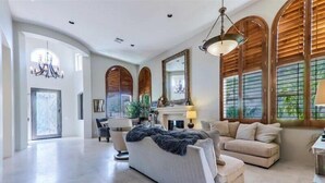 A spacious living room with a large sofa, a smaller armchair, and a coffee table. There is a fireplace with a mirror above it and a chandelier hanging from the ceiling.