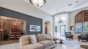 A spacious living room with a beige sofa, a coffee table, and a fireplace. There is a large window with blinds and a chandelier.