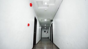 Un long couloir étroit aux murs blancs et au sol ciré. Des portes se trouvent de part et d’autre, et un détecteur d’incendie est fixé au mur gauche.