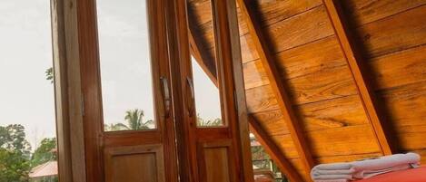 Una habitación con techo de madera, una cama con manta roja y una ventana con vista a la vegetación.