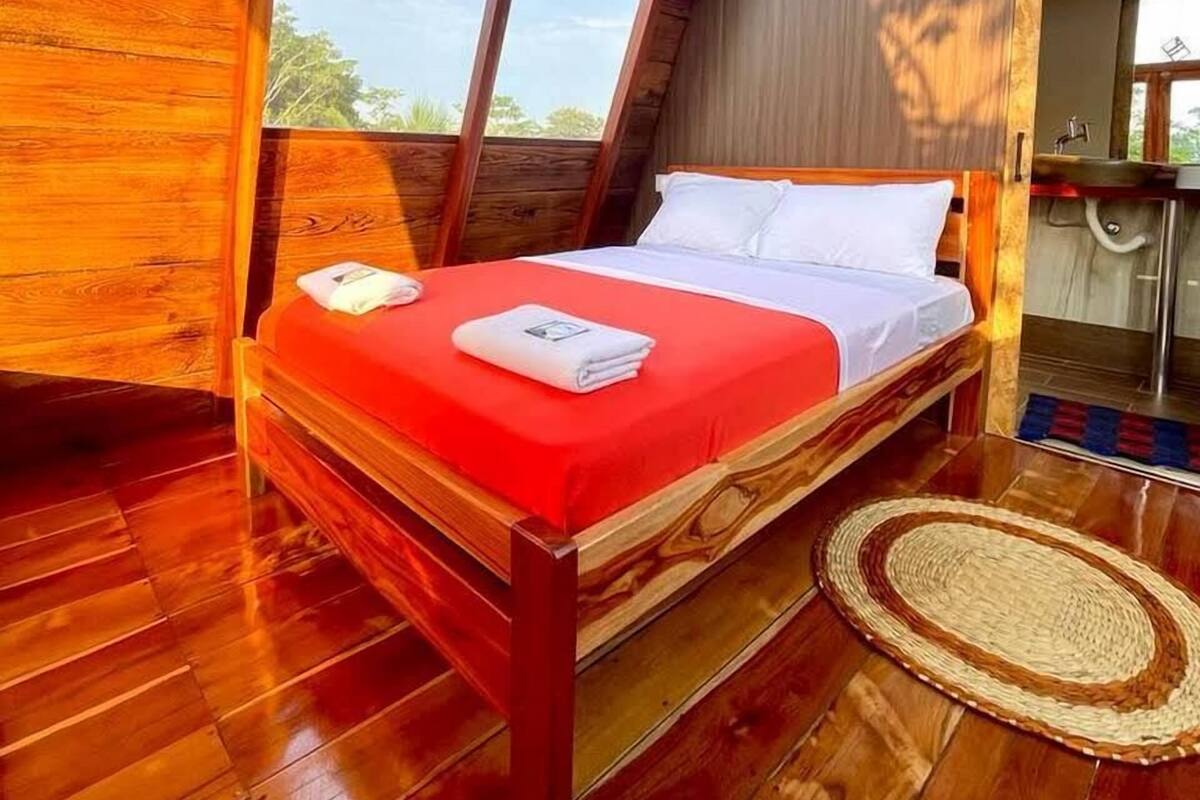 Una cama con estructura de madera, cubierta con una colcha roja y almohadas blancas, en una habitación con piso de madera y un ventanal grande.