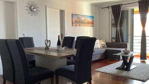 Een eetgedeelte met een houten tafel en donker beklede stoelen, een stadsgezichtschilderij aan de muur, en een bank op de achtergrond.