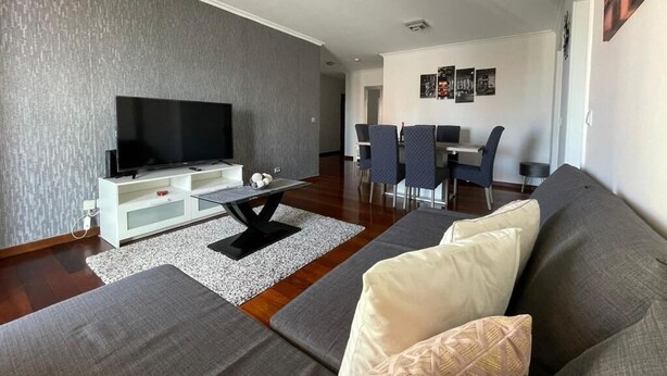 Een moderne woonkamer met een flatscreen televisie, een grijze bank en een eethoek met stoelen en een tafel.