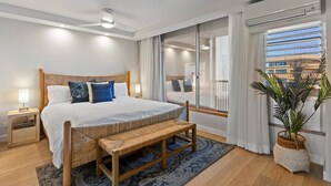 Ein modernes Schlafzimmer mit einem großen Bett, Nachttischen, einer Bank, einer Topfpflanze und Blick auf die Stadt durch große Fenster.