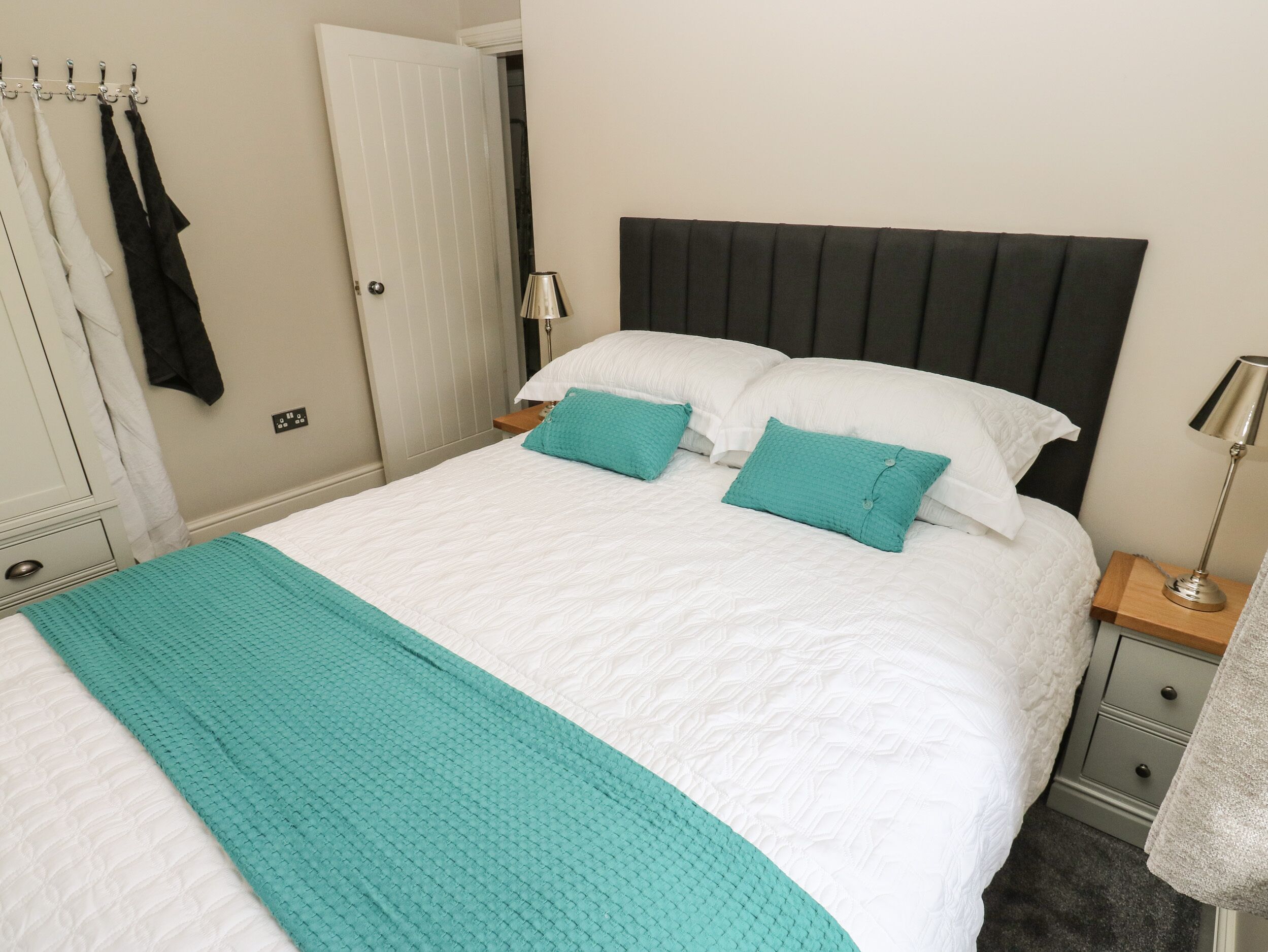A neatly made bed with a teal throw blanket and pillows, a bedside table with a lamp, and a wardrobe with hanging clothes.