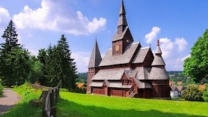 Une église traditionnelle, dotée d’un toit à forte pente et de plusieurs flèches, entourée d’une végétation luxuriante et surplombée d’un ciel bleu clair.