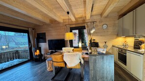 L’intérieur chaleureux d’un chalet avec un plafond en bois, un coin repas comprenant une table et des chaises, une cuisine équipée d’appareils modernes et une grande fenêtre offrant une vue sur l’extérieur.