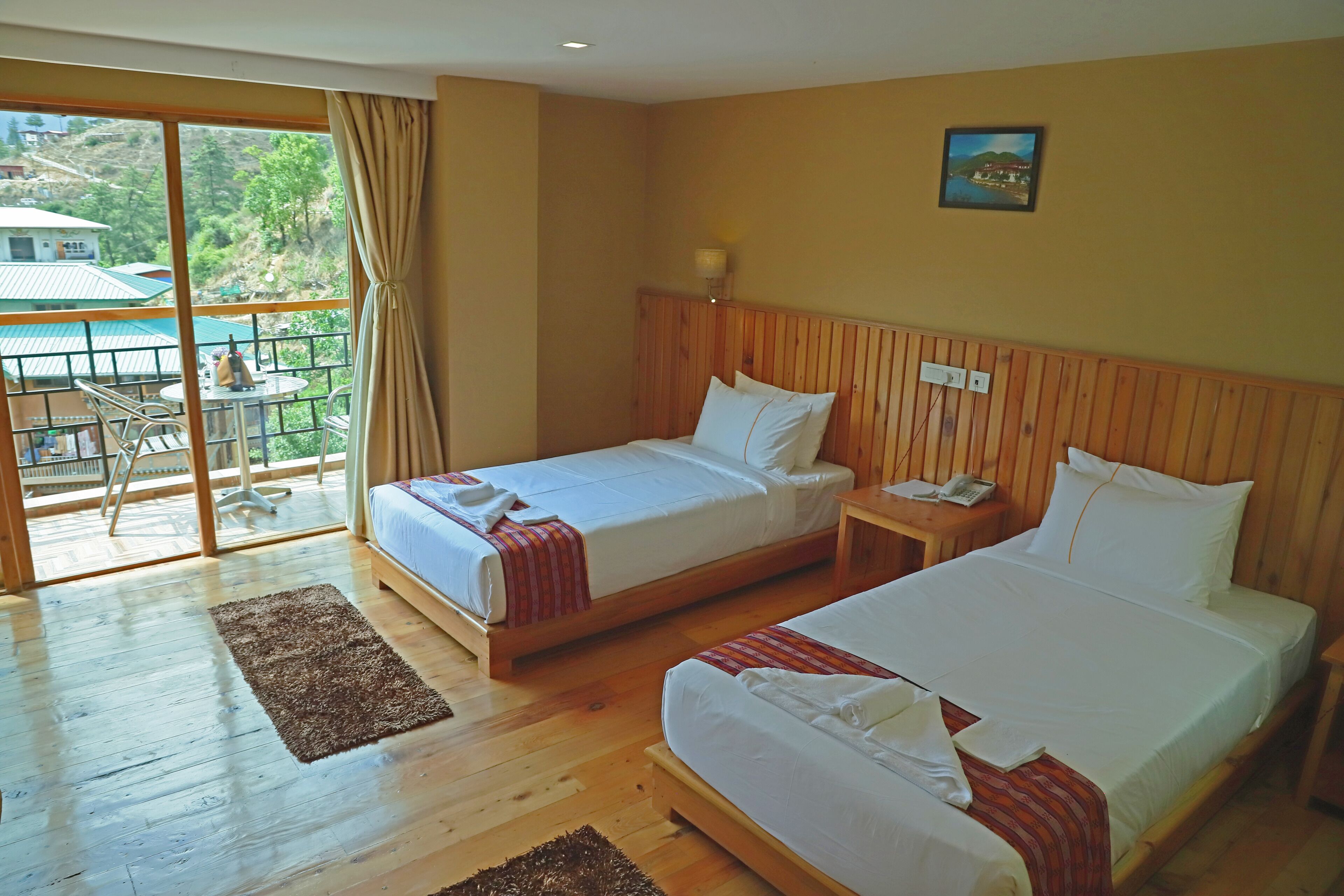 Hotelkamer met twee bedden, een houten hoofdeinde, een tafeltje met een telefoon en uitzicht op een balkon met tuinmeubilair.