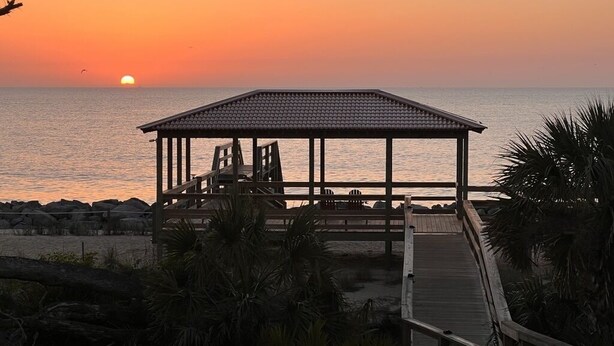 Un quiosco frente a la playa con piso de madera y zona de estar, con vista al océano al atardecer.
