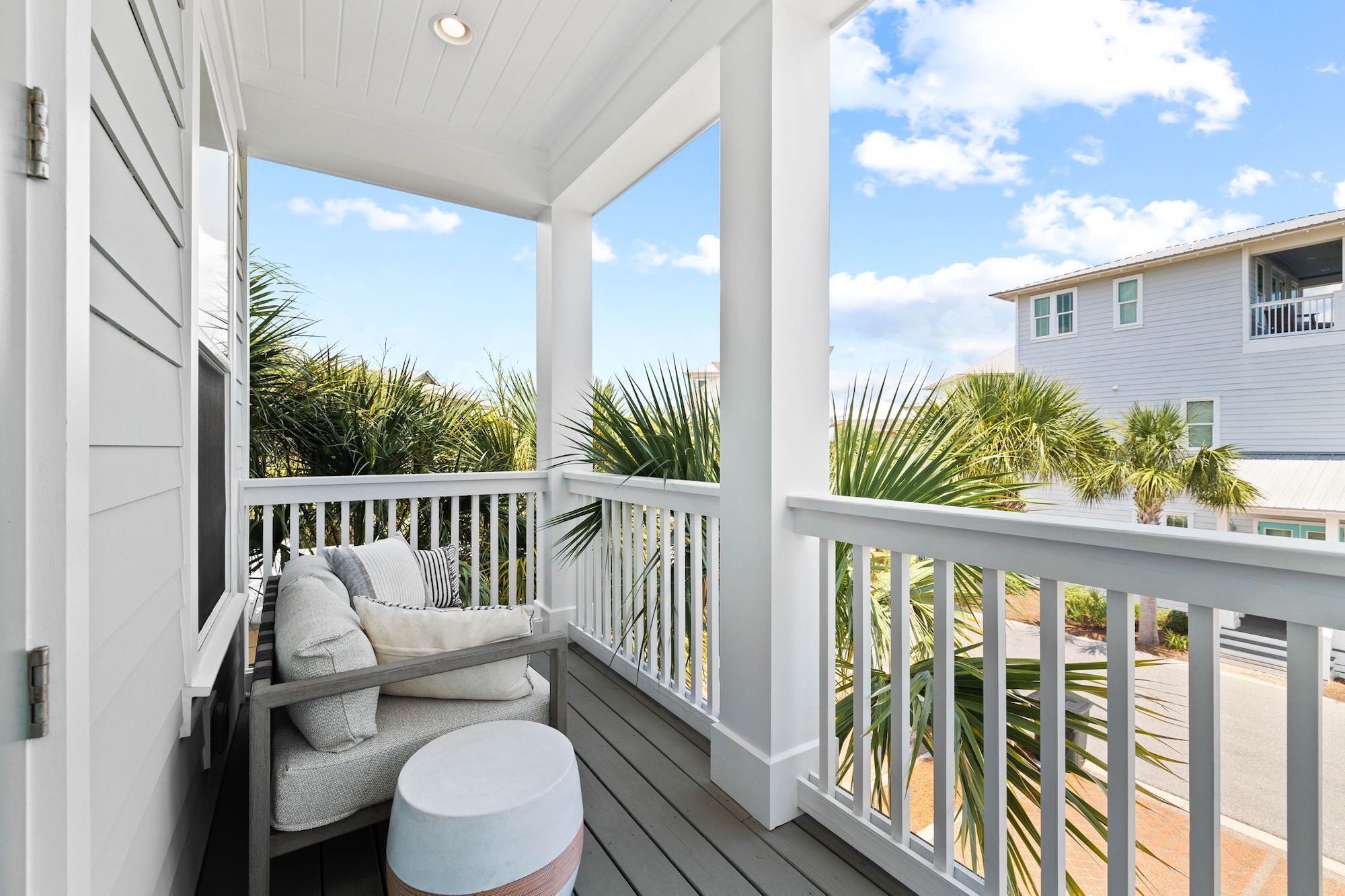 A covered porch with a cushioned seating area and a round table, overlooking a residential street with palm trees and a house.
