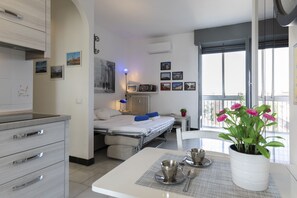 Una habitación de hotel moderna con kitchenette, un comedor con mesa para dos, una cama con vistas y un balcón lleno de flores.