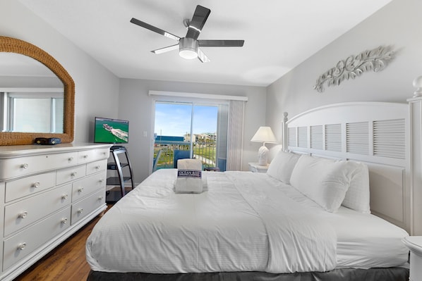 A hotel room with a bed, a ceiling fan, a dresser, a television, and a view of the ocean.