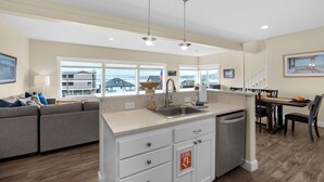 A modern kitchen with a central island, a dining area with a table and chairs, a sofa, and a view of the outside. - SUNSET DREAMS at Seaside Beach (Seaside)