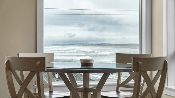 A dining table with four chairs, a glass top, and a view of the ocean through a window. - SUNSET DREAMS at Seaside Beach (Seaside)