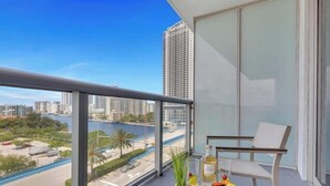 A balcony with a view of a cityscape and water, a chair, and a table with a bottle and fruit. - Sunset Oasis: 2BR, Balcony, Pool, Perfect Getaway (Hallandale Beach)