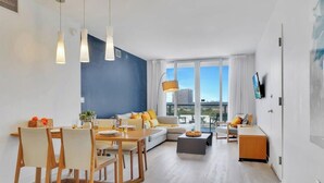 A modern living room with a dining area, a sofa set, a coffee table, and a TV mounted on the wall. - Sunset Oasis: 2BR, Balcony, Pool, Perfect Getaway (Hallandale Beach)