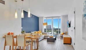 A modern living room with a dining area, a sofa set, a coffee table, and a TV mounted on the wall. - Sunset Oasis: 2BR, Balcony, Pool, Perfect Getaway (Hallandale Beach)