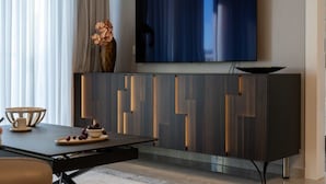 A modern living room with a flat-screen TV mounted on the wall, a dark wooden cabinet with geometric cutouts, a dining table with a bowl and cherries, and a vase with flowers on the cabinet. - Enchanting 2 Bedroom In Best Location/Parking (Jerusalem)