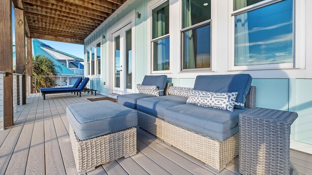 A covered outdoor seating area with wicker furniture and cushions, large windows, and a view of a blue building in the background.