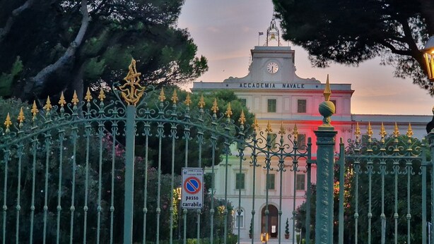 Una entrada protegida por una reja ornamentada, una torre del reloj y un cartel que dice 'Accademia Navale'.