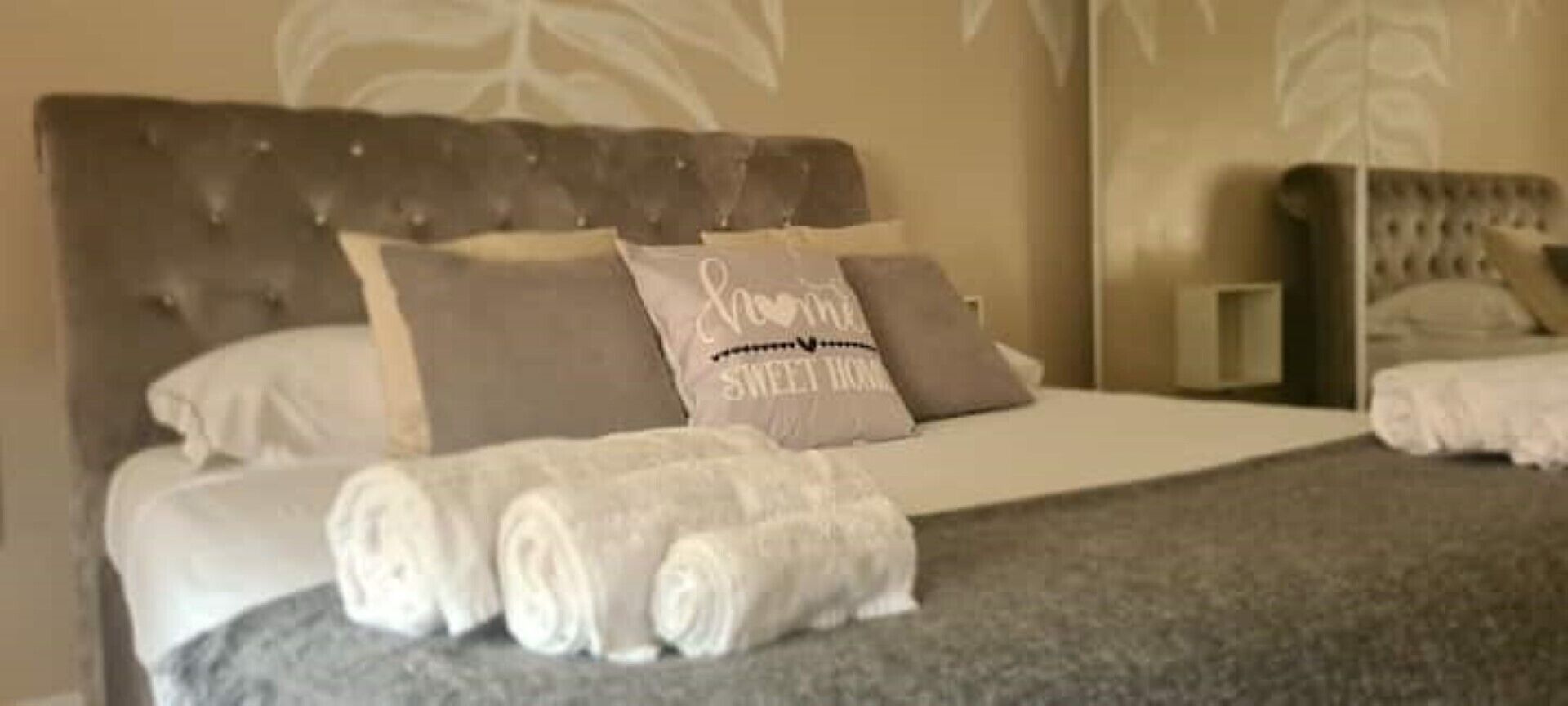 Een net opgemaakt bed met opgerolde handdoeken en een kussen met de tekst 'Sweet Home'.
