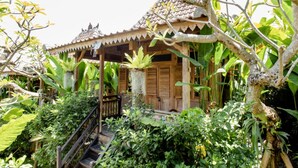 Une maison traditionnelle en bois, avec un toit en tuiles, entourée d’une végétation luxuriante et un escalier menant à l’entrée.