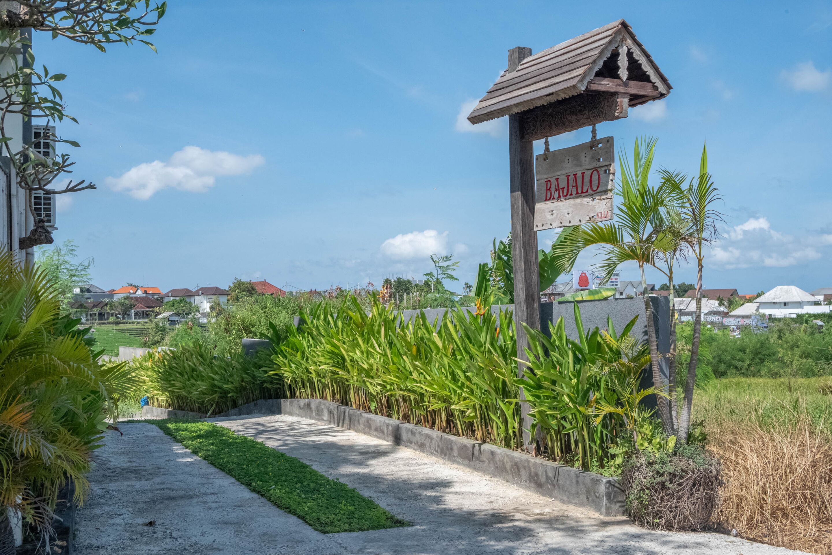 A pathway leading to a wooden signpost with 'BAJALO' written on it, surrounded by lush greenery and a clear sky.