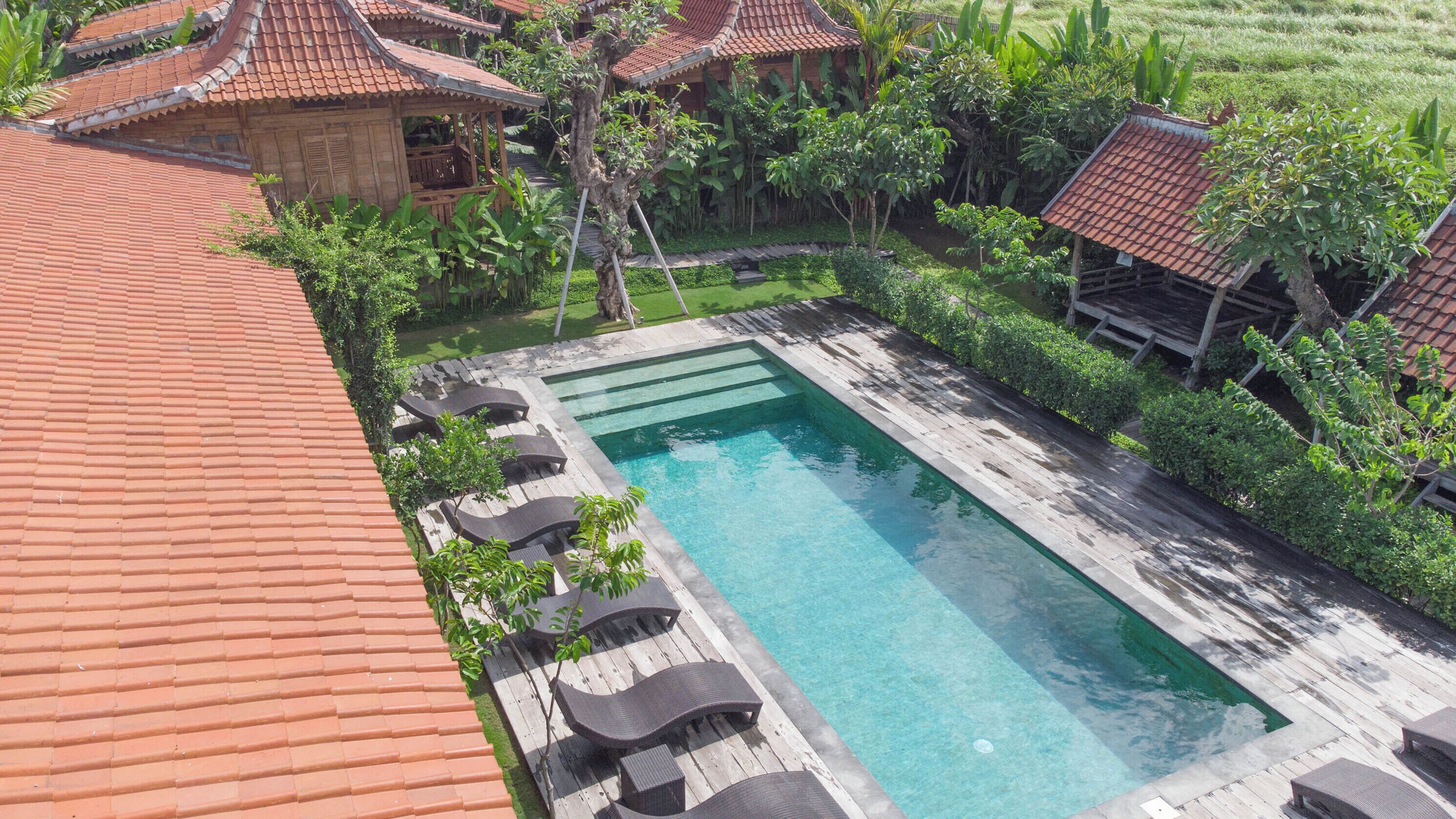 A rectangular swimming pool with a wooden deck and lounge chairs, surrounded by lush greenery and traditional buildings with red-tiled roofs.
