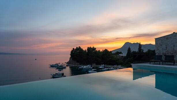 Una vista tranquilla della piscina con barche ormeggiate vicino a una scogliera, con vista su un mare calmo e un tramonto.