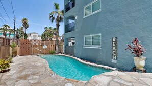 A pool area with a stone patio, a wooden fence, and a building with a 'BEACH' sign. - Gulf Coast Condominium Unit 4 2 Bedroom Condo by RedAwning (South Padre Island)