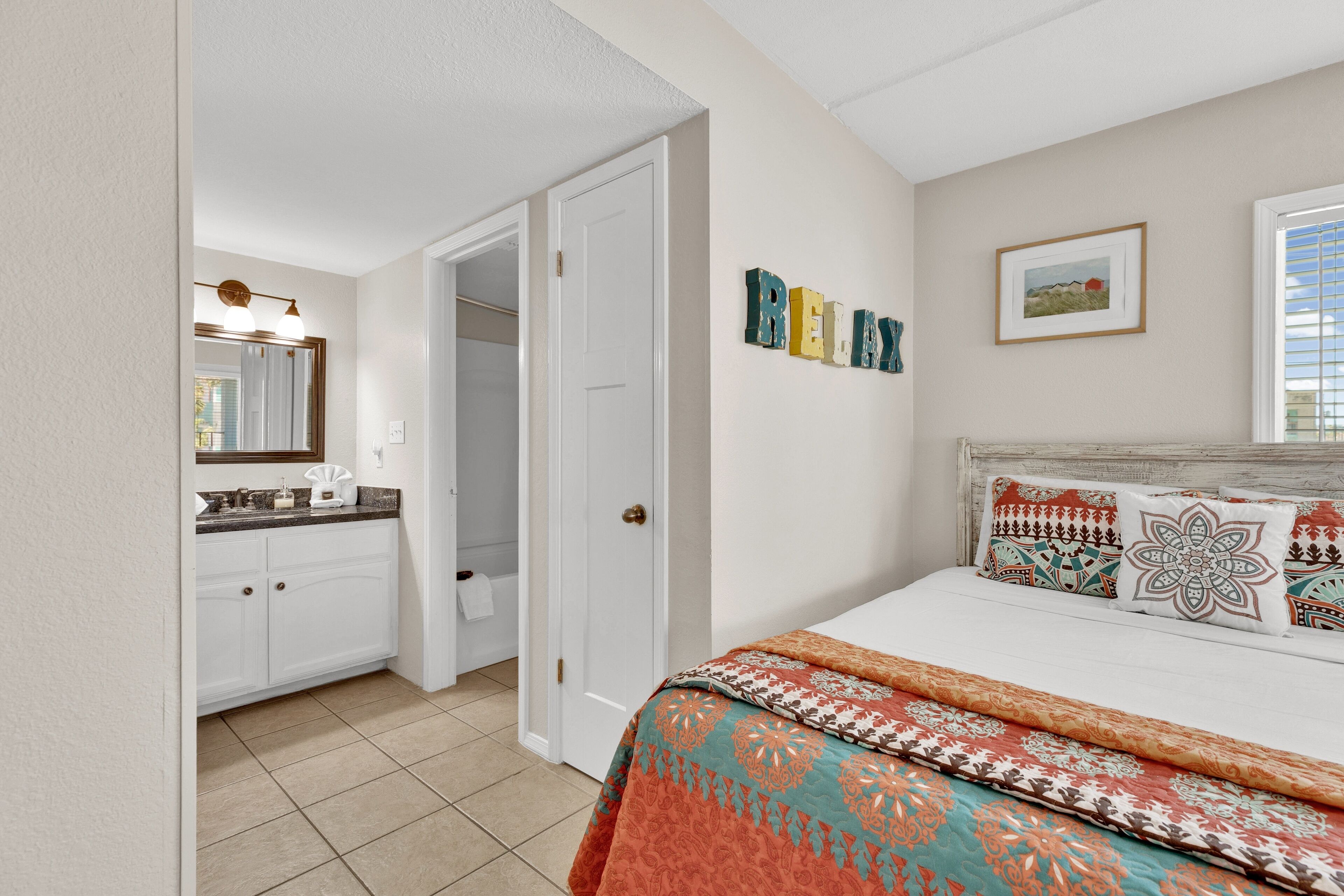 A bedroom with a bed, a bathroom with a sink and mirror, and a door leading to another room.
