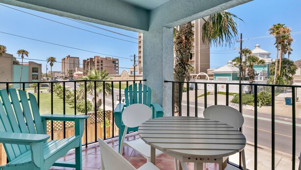 A balcony with teal chairs and a table, overlooking a street with buildings and palm trees. - Gulf Coast Condominium Unit 4 2 Bedroom Condo by RedAwning (South Padre Island)
