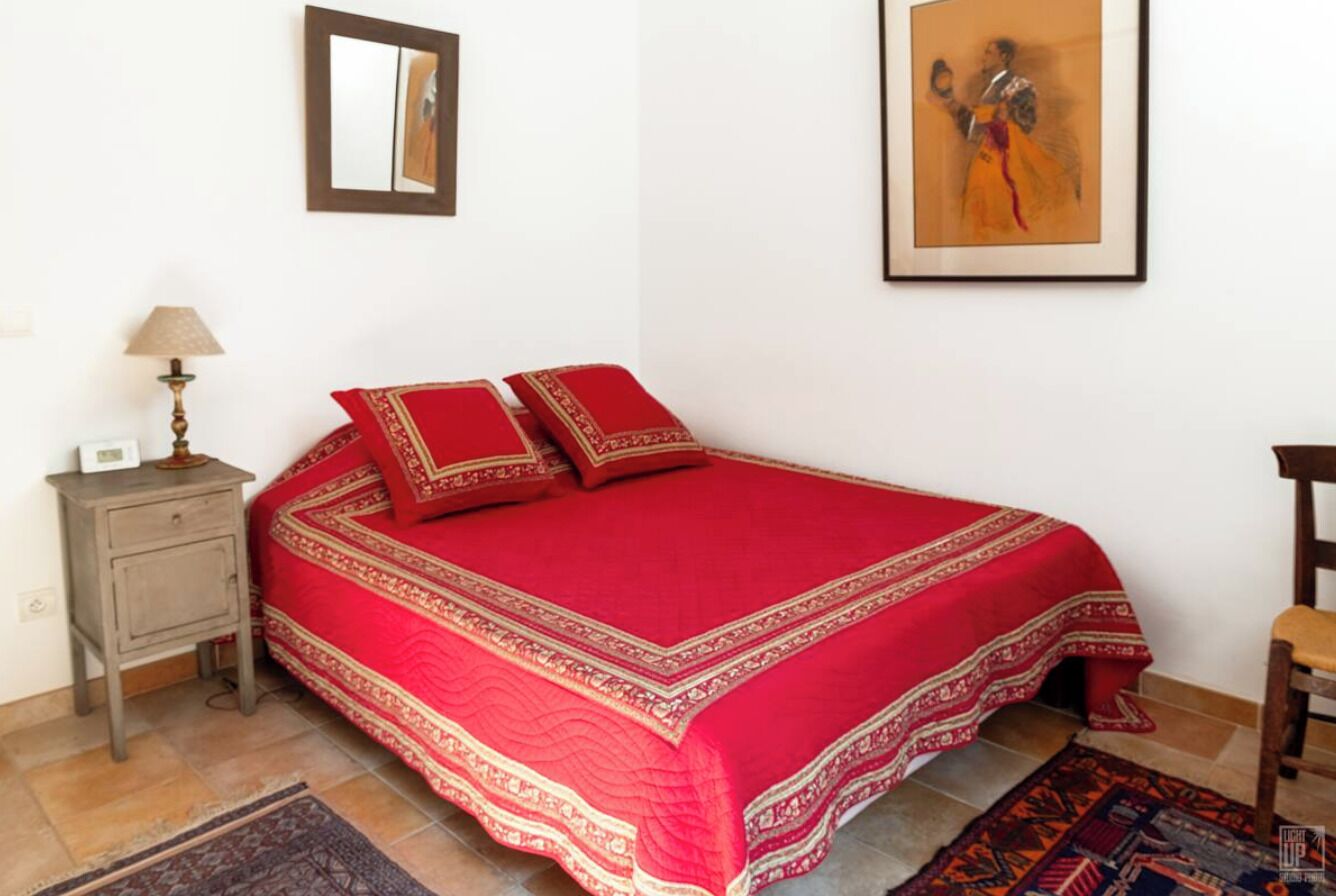 Een netjes opgemaakt bed met een rode sprei en kussens, een klein nachtkastje met een lamp, en een ingelijst beeld aan de muur.