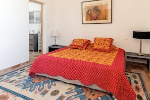 A bedroom with a bed, a nightstand, a lamp, a framed picture on the wall, and a view of the bathroom.