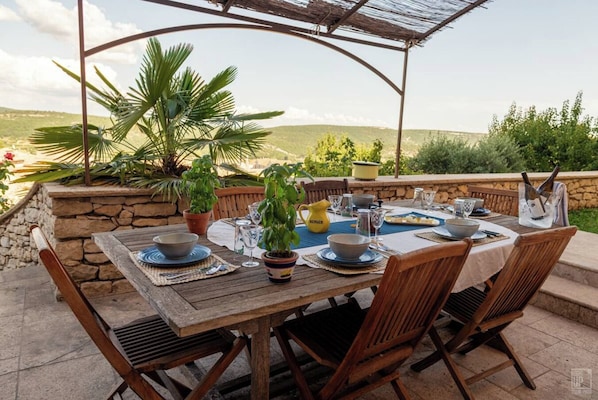 An outdoor dining area with a wooden table set for four, surrounded by stone walls and potted plants.