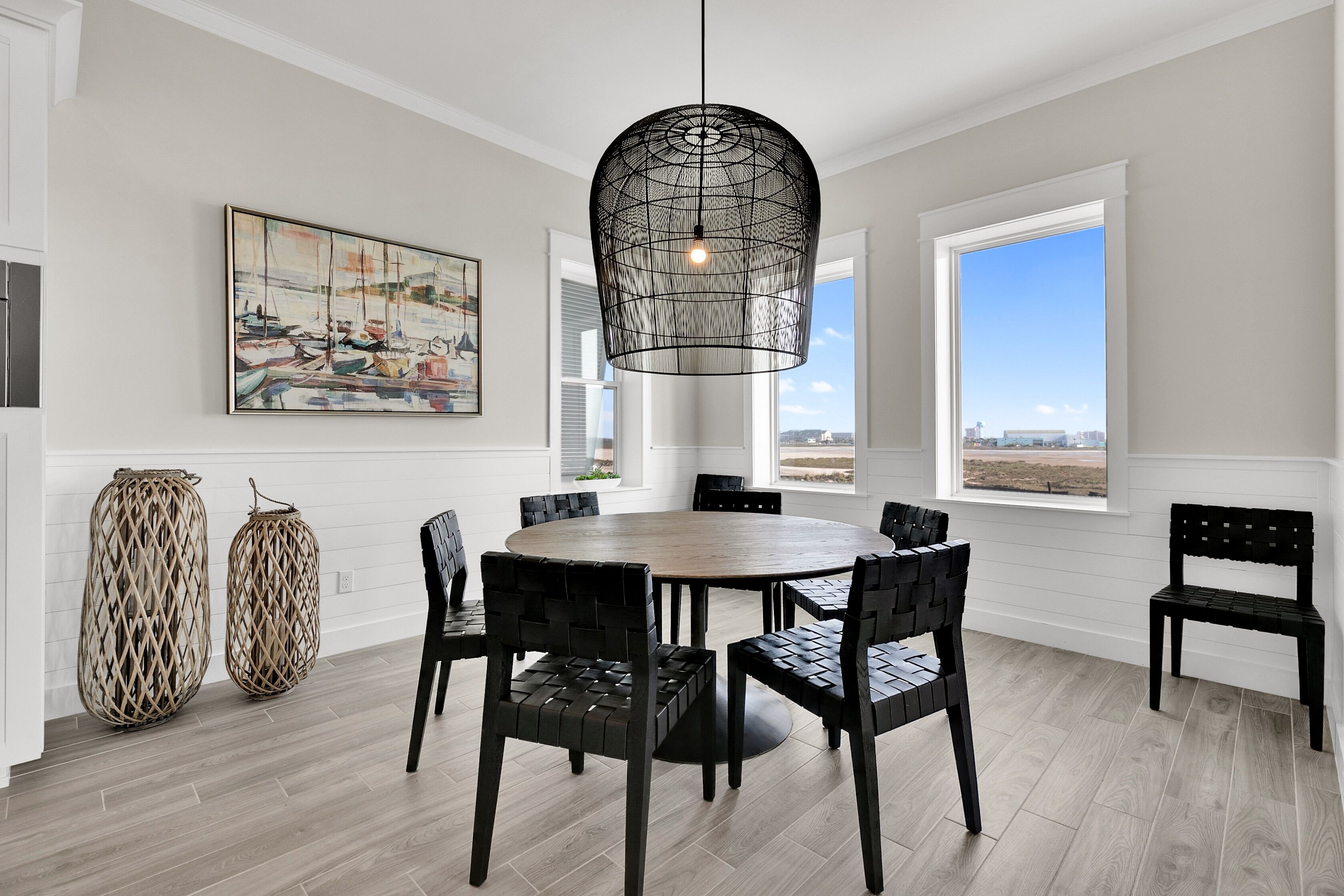 Een eetruimte met een ronde tafel, zwarte stoelen, een geweven hanglamp en een groot raam met uitzicht.