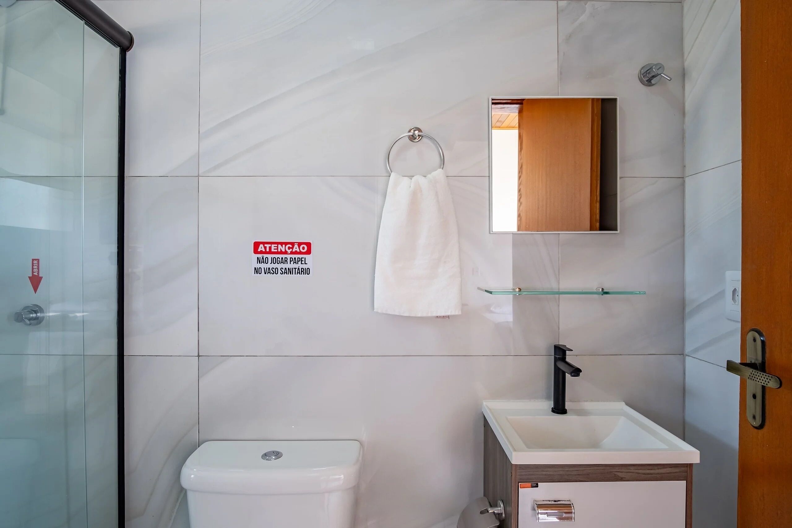 Baño con inodoro, lavamanos y cabina de ducha de vidrio. Una toalla cuelga de un aro metálico, y un espejo está colgado sobre el lavamanos.