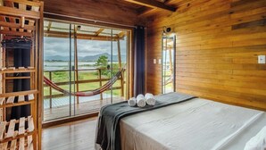 A wooden cabin room with a bed, a hammock, and a view of a lake and mountains. - Bangalos Luz da Lagoa (Imbituba)