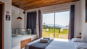 A bedroom with a large window, a bed, a wooden ceiling, and a view of a green landscape. - Bangalos Luz da Lagoa (Imbituba)