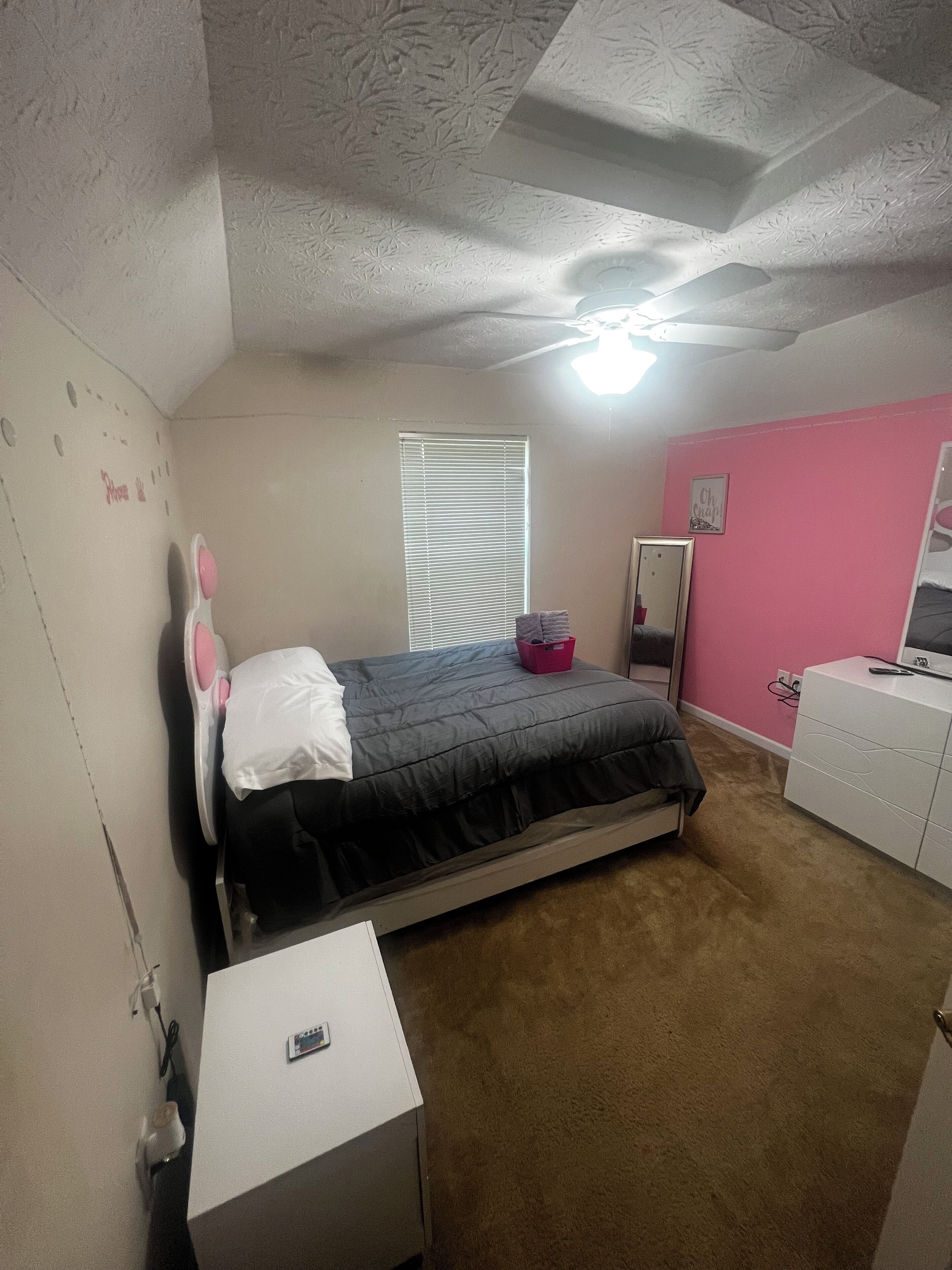 Un dormitorio con cama, cómoda, espejo, ventana con persianas y ventilador de techo.