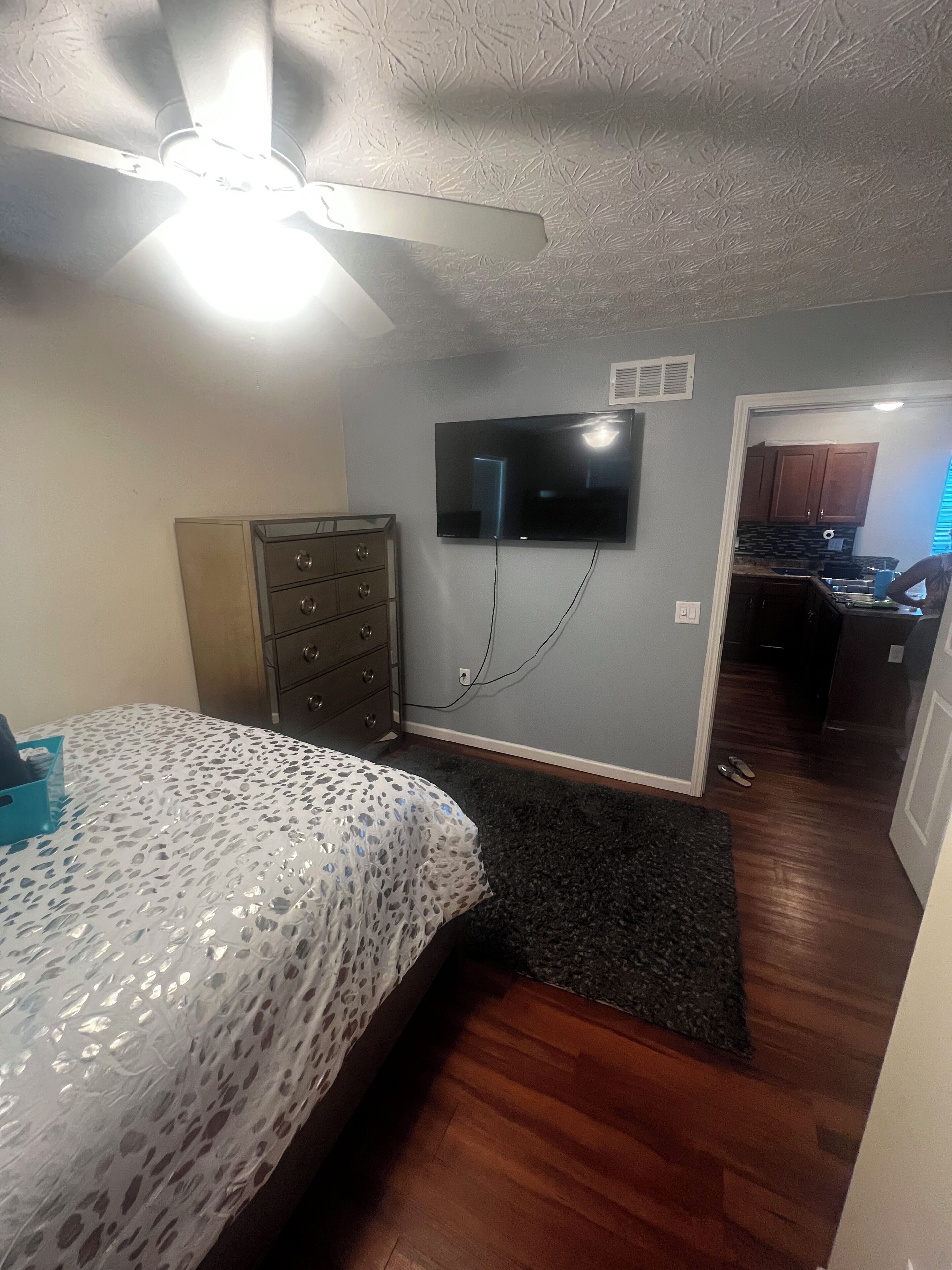 A bedroom with a bed, dresser, ceiling fan, and a TV mounted on the wall.